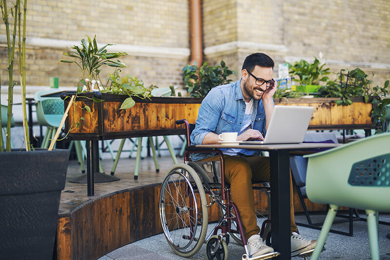 Man in wheelchair is sitting at a table while using a laptop while on the phone.